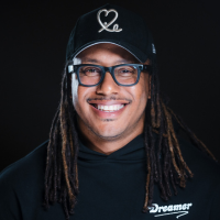 Smiling man with dreadlocks wearing a "Dreamer" hoodie and a baseball cap.